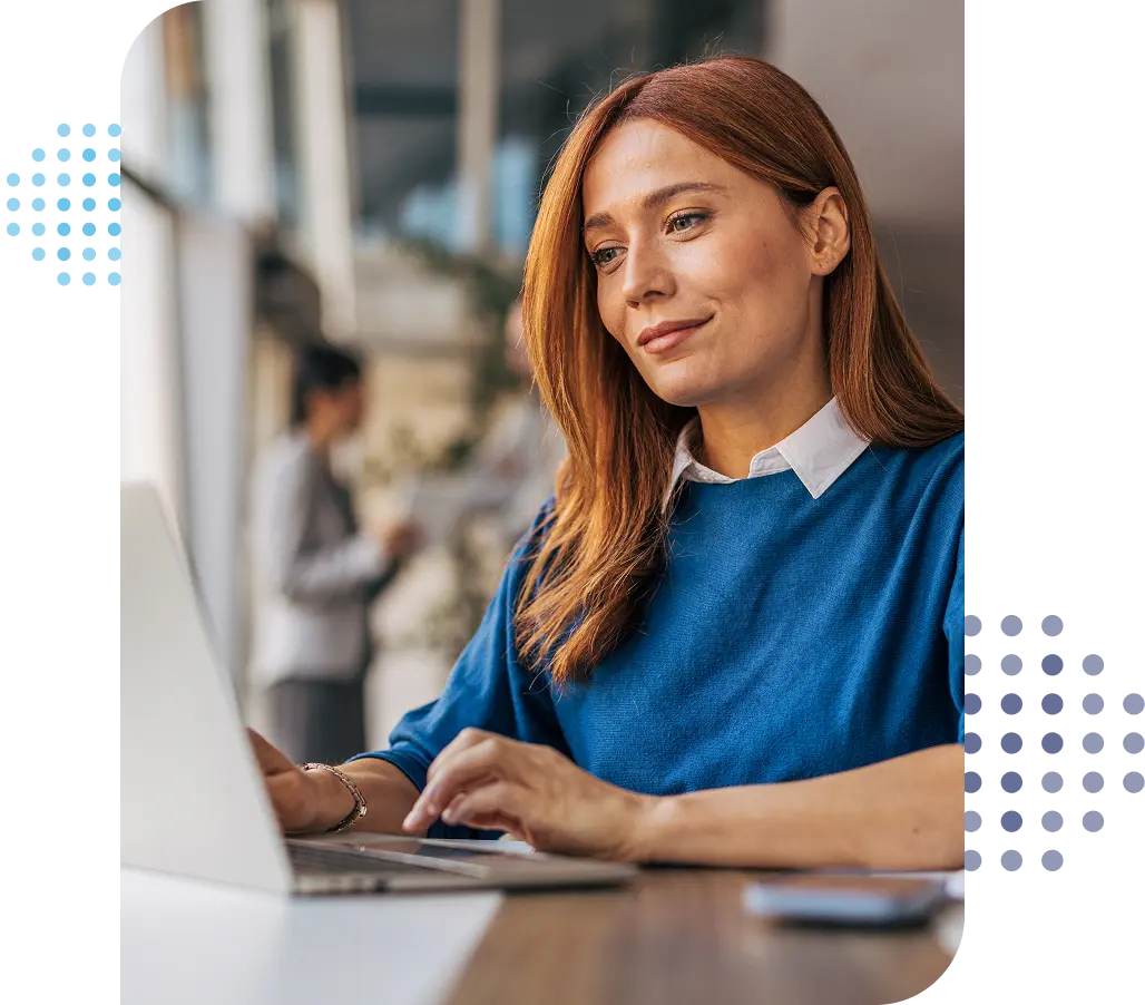 smiling woman in a button down shirt with a blue sweater over it sitting at a table looking at a laptop
