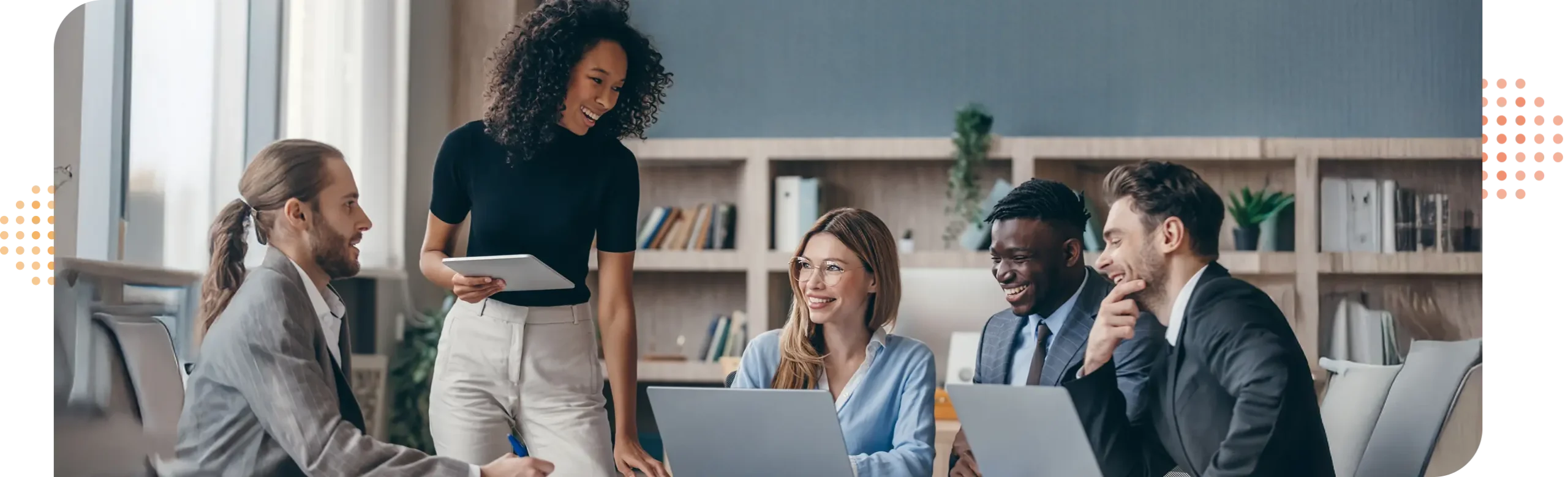 people in business attire sitting at a meeting table smiling & talking while looking at a laptops