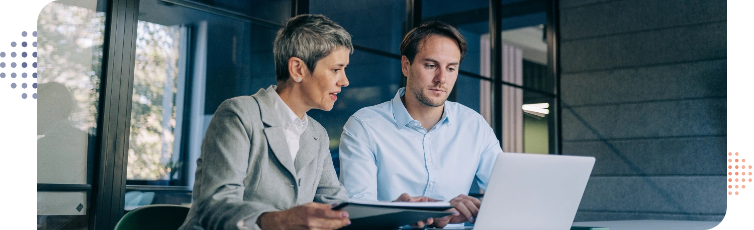 man & woman in business casual sitting at a table looking at a laptop & papers